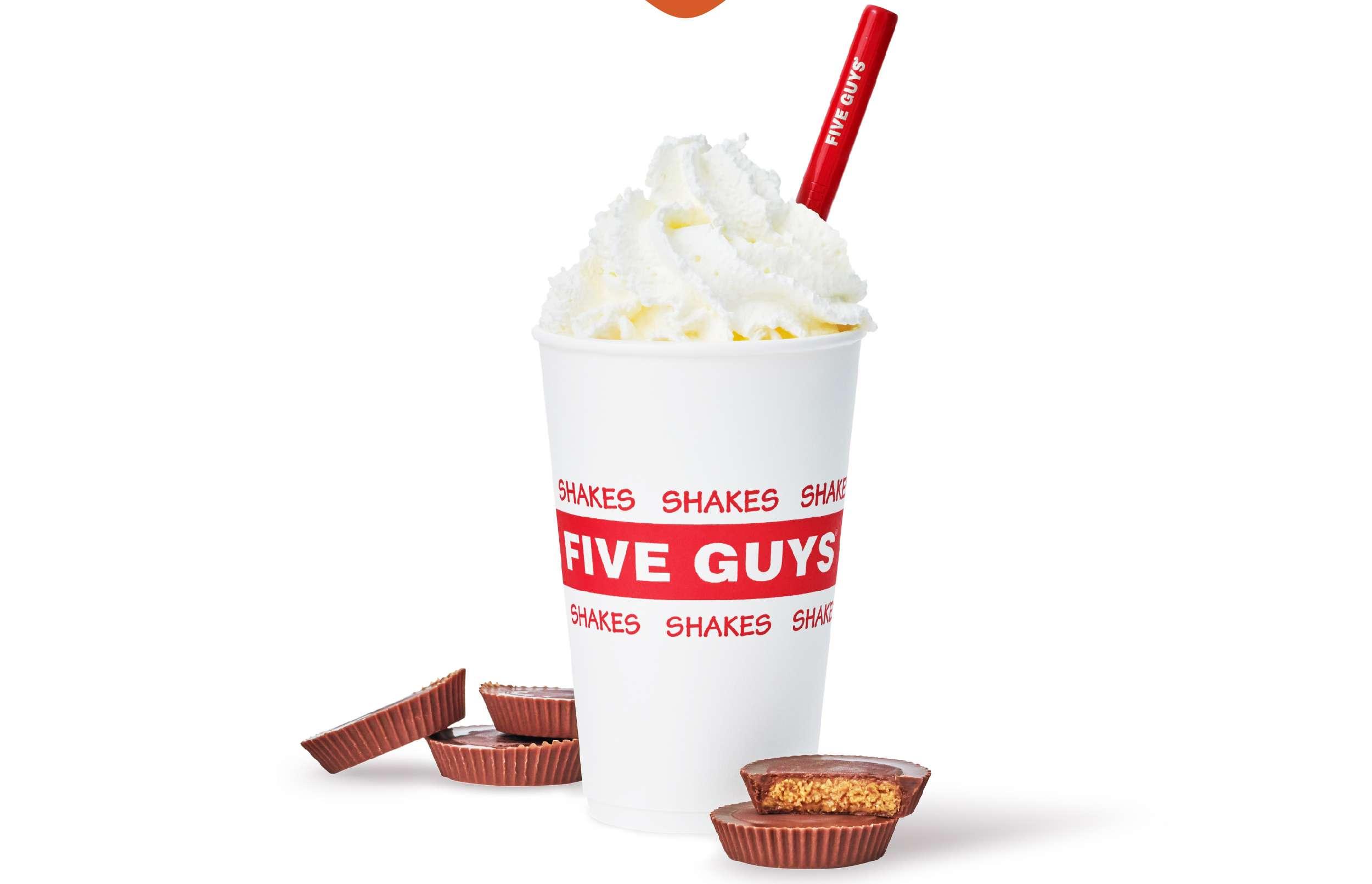 A Five Guys milkshake in a white cup with red "FIVE GUYS SHAKES" branding, topped with whipped cream and a red straw. Surrounded by several peanut butter cups, one of which is cut in half to show the filling.