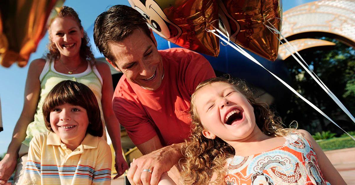 A joyful family of four smiling and laughing together, with the young girl in the foreground laughing and holding character balloons, under a sunny sky at what appears to be a theme park.