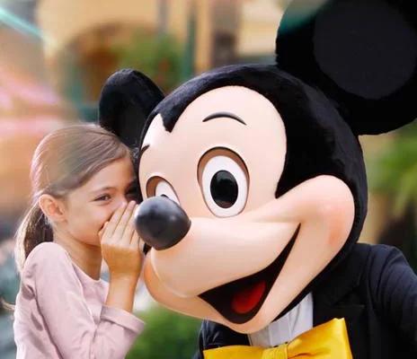 A young girl whispering into the ear of a person in a Mickey Mouse costume, both smiling and standing in a colorful outdoor setting.