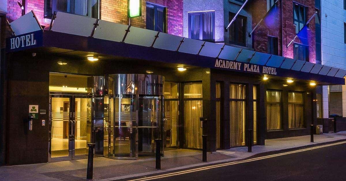 Exterior view of the Academy Plaza Hotel at night. The entrance is brightly lit with glass doors, including a revolving door, and warm interior lighting visible inside. The hotel name is displayed on a dark awning above the entrance. The building facade features brick and modern elements, with purple and blue accent lighting on the upper floors.