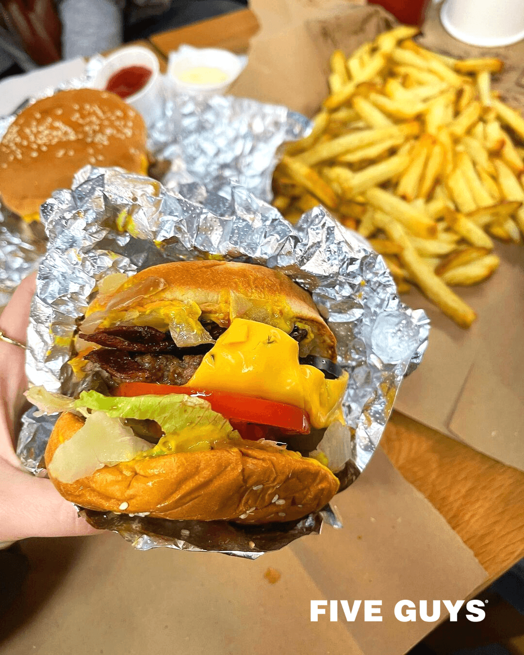 A close-up shot of a juicy cheeseburger from Five Guys wrapped in foil, held in hand. The burger is packed with vibrant toppings including melted cheddar cheese, crispy bacon, lettuce, tomato, pickles, onions, and mustard. In the background, a generous serving of golden fries and another burger are visible on a brown paper-lined tray. The signature FIVE GUYS logo is displayed in the bottom right corner.